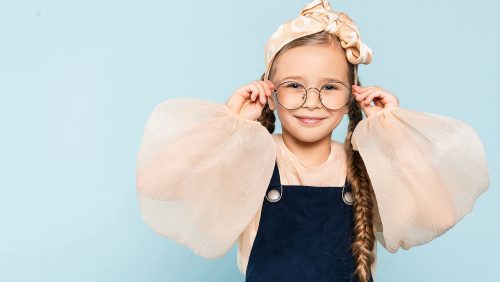 cheerful kid adjusting glasses while looking at camera isolated on blue