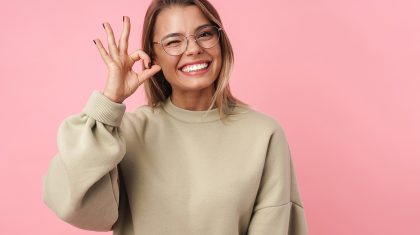 Portrait of young smiling woman in eyeglasses winking and gesturing ok sign isolated over pink background